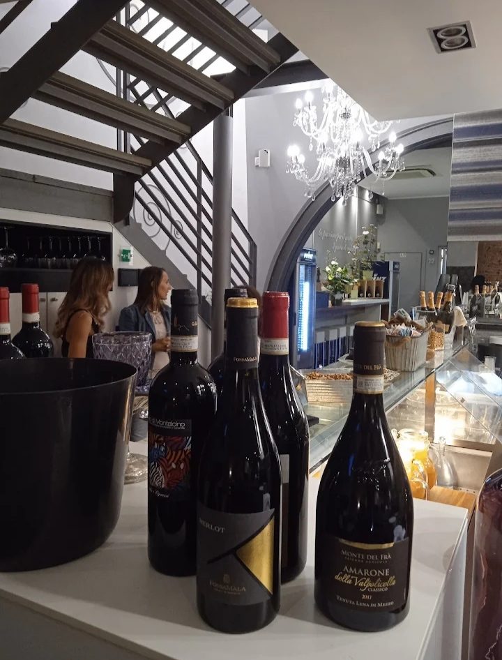 Bottiglie di vino rosso allineate sul bancone di MAMA'S: Brunello di Montalcino, un Merlot e un Amarone della Valpolicella, con la vetrina dei dolci e la scala sullo sfondo.