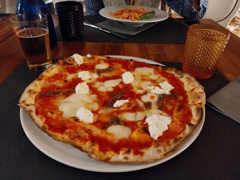 Pizza rossa con fiocchi di burrata e acciughe su un tavolo apparecchiato, con i bicchieri e un piatto di pasta sullo sfondo.