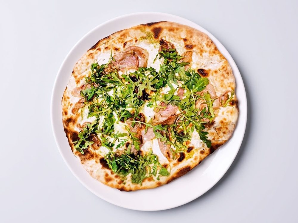 Pizza bianca vista dall'alto con prosciutto crudo, stracchino e una manciata di rucola, su un piatto bianco.