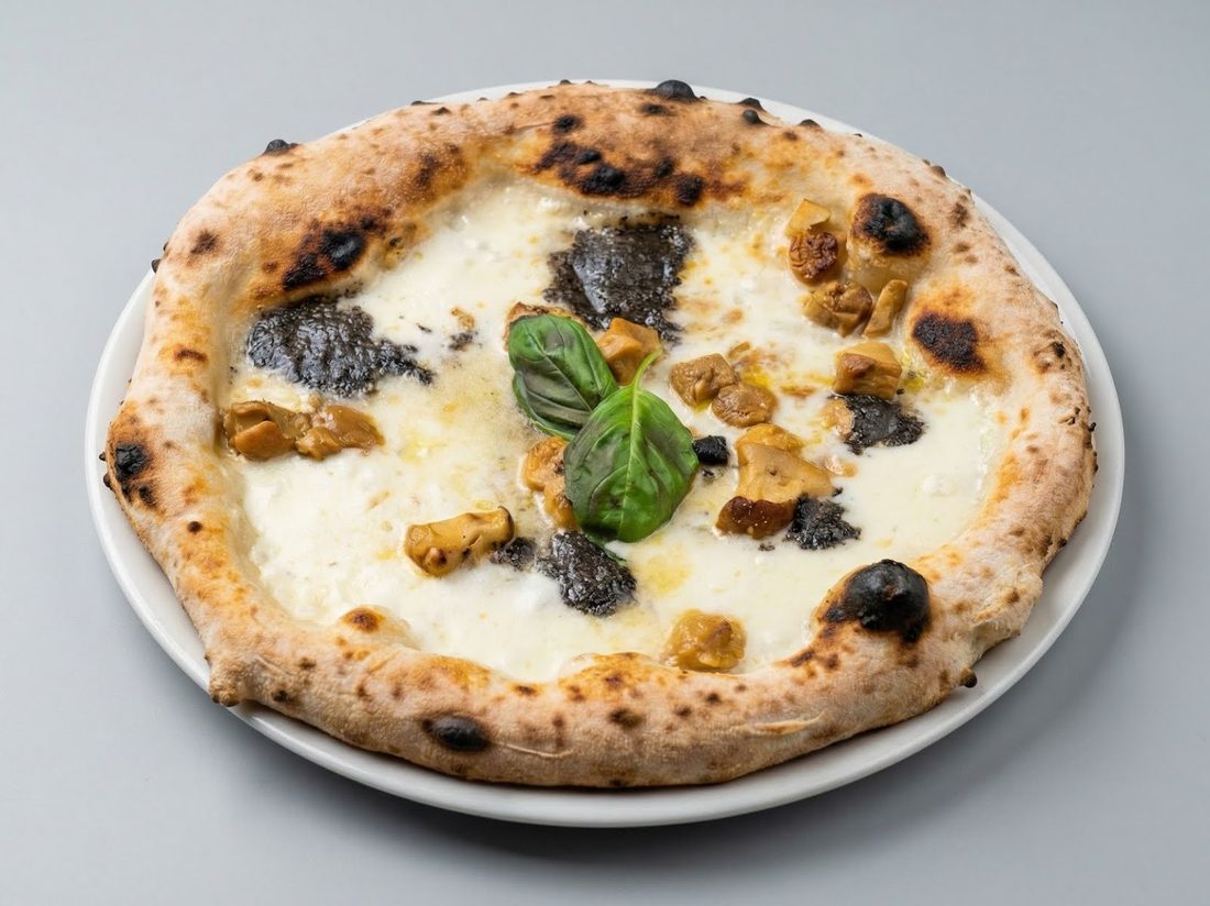 Pizza bianca con porcini a pezzi, crema di tartufo nero e una foglia di basilico al centro, servita su un piatto bianco.