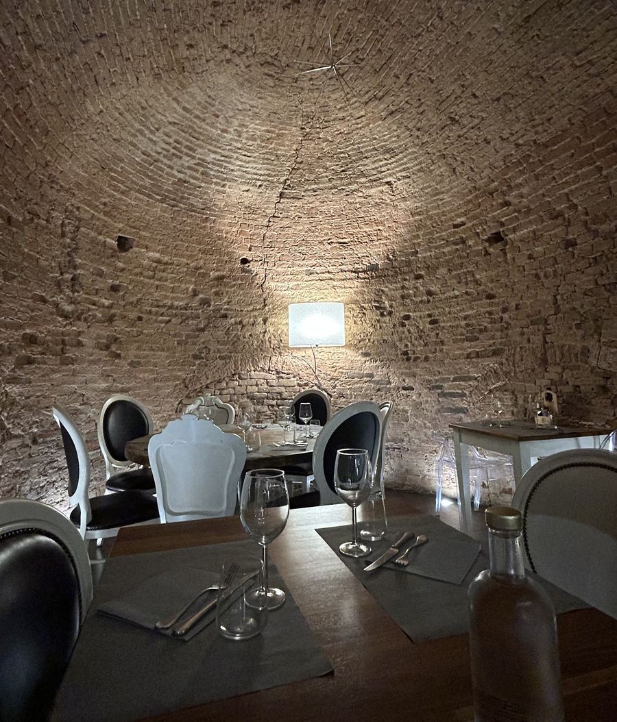 La sala di MAMA'S sotto una volta a botte in mattoni a vista, con tavoli apparecchiati, sedie bianche a medaglione e una lampada a parete che illumina il fondo.