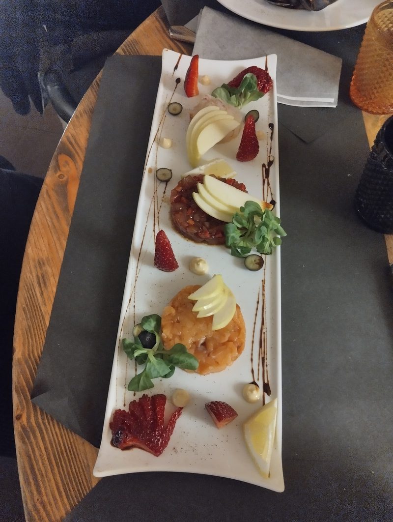 Tre tartare servite su un piatto lungo bianco, con fettine di mela, insalatina, fragole e gocce di salsa.