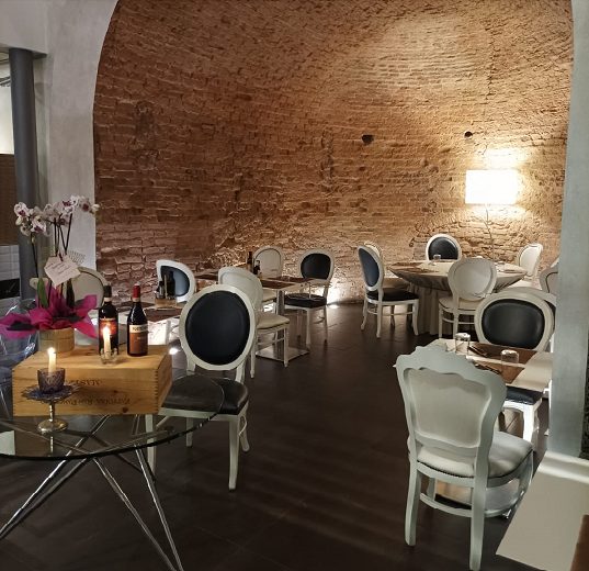 La sala sotto la volta in mattoni: sedie bianche a medaglione con la seduta scura, tavoli apparecchiati e un mazzo di orchidee su un tavolo di vetro in primo piano.